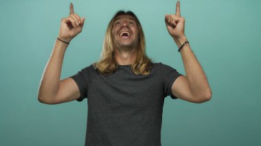 Sarışın, uzun saçlı bir adam, gümüş rengi stüdyo ortamında gülümserken iki işaret parmağını da yukarı kaldırıyor. İyimserlik umudu..