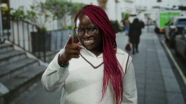 Kırmızı örgülü, gözlüklü, gülümseyen ve dışarıdaki bir caddede kameraya bakan bir kadın..