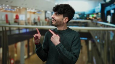 Genç adam, hareketli bir alışveriş merkezini işaret ederek canlı bir alışveriş havası ve şaşkınlık ve merak ifadeleri sergiliyor..