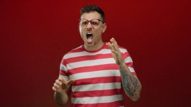 Dövmeli, gözlüklü, çizgili tişörtlü, kırmızı arka planda duygularını ifade eden bir adam..