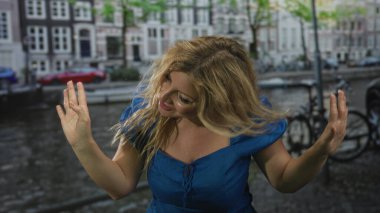 Elleri havada olan bir kadın mavi elbiseli bir su kenarında avuçlarını açıp şarkı söyleyen bir kadın..