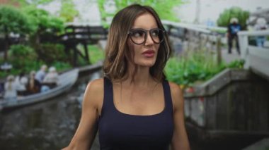 Gözlüklü ve mavi kolsuz tişörtlü genç bir kadın, sokakta parmağını kaldırıyor..