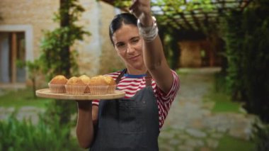 Kadın, taş yol ve yeşillik girişinde ahşap kek tepsisi tutuyor ve kamerayı işaret ediyor; misafirperverliği davet ediyor..
