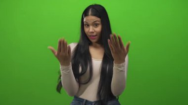 Parlak yeşil stüdyoda bej rengi bluzla gülümseyen genç Latin kadın avuçlarını açık gösteriyor; belirsizlik..