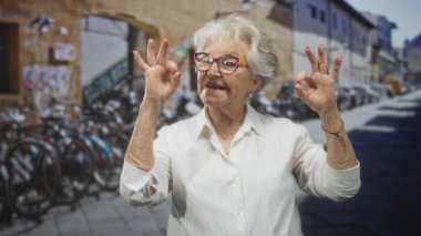 Kadın, park halindeki bisiklet ve eski binalarla dolu bir caddede elleri ve renkli gözlükleriyle tamam işareti gösteriyor..