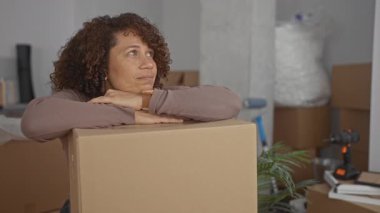 Yeni binanın içinde kollarını kavuşturmuş bir kadın, boş odada sakin bir ifadeyle yukarıya bakıyor; beklenti.