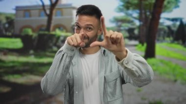 İspanyol adam ezik hareketler yapıyor ve doğayla çevrili yeşil güneşli parkta kamerayı işaret ediyor.
