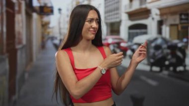 Bir şehir caddesinde kırmızı bir tişörtle çıplak omuzlarını tararken yüzünü buruşturan kadın, mağaza vitrinleri ve scooterlar arkasında bulanıklaştı;.