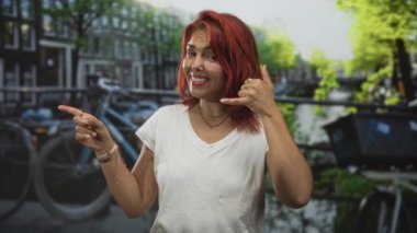 Sokakta kadın sol eliyle işaret ediyor ve sağ eliyle çağrı hareketi yapıyor, gülümsüyor ve beyaz tişört ve kolye takıyor; oyuncu özgüven.