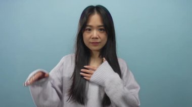 Genç Çinli bir kadın mavi arka plana karşı yeminli bir jest yapıyor, rahat bir süveter giyiyor, kendinden emin bir ifadeyle ileriye bakıyor, umut vadeden bir konsepti net bir şekilde gösteriyor..