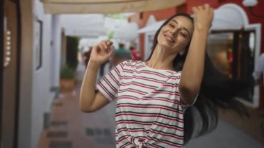 Gülümseyen ve elini saçlarını tarayan bir kadın işlek bir yaya caddesinde dükkanlar ve brandalarla kaplı, çizgili bir düğümlü tişört giyen, kaygısız bir an.