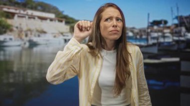 Stüdyo limanında eli kulağında ve yumruğunu kaldırmış bir kadın demirli tekneler ve ahşap rıhtımla birlikte..