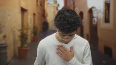 Dar kaldırım taşı caddesindeki yıpranmış binaların yanında gözlüklü ve beyaz kazaklı genç adam..