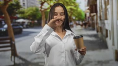 Meşgul bir caddede beyaz gömlekle yürürken elinde kahve fincanı tutan kadın tamam işareti gösteriyor, neşeli bir onay..