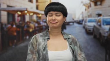 Dışarıda, kalabalık bir caddede terasta neşeli bir ifadeyle bir kadın, arabalarla, koruma jestleri sergiliyor ve açık bir şehir restoranında katmanlı kıyafetler giyiyor..