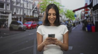 Meşgul bir şehir caddesinde, park halindeki arabalar ve ağaçlarla akıllı telefon tutarken gülümseyen genç İspanyol kadın, ekrandan aşağıya bakıyor;.