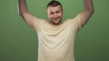 Çıplak kollu bir adam stüdyoda yeşil bir duvara karşı ellerini kaldırıyor, kapalı gözlerle gülümsüyor; neşeli bir rahatlık.