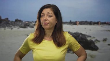 Elleri kalçalarının üzerinde, açık gökyüzünün altında, kayalık bir sahil sahilinde, üzerinde sarı gömlek olan bir kadın, sessiz bir tefekkür..