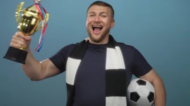 Elinde kupa ve futbol topu tutan adam mavi stüdyoda siyah beyaz atkı takarak bağırıyor..
