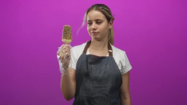 Stüdyoda elleri eldivenli, çikolata kaplı buzlu dondurma tutan, mor arka plan ve kot önlüklü bir kadın..
