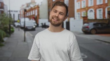 Sakallı ve beyaz tişörtlü bir adam dil uzatıyor ve başını eğiyor, geniş bir gülümseme ile görünür yüz ve boyun, park etmiş arabalar ve tuğla binalar ile bir şehir caddesinde duruyor; neşeli gençlik yaramazlık.