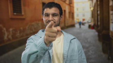 Gözlüklü ve kot ceketli genç İspanyol adam yıpranmış binalarla dolu dar bir eski kasaba sokağında dikilirken kamerayı işaret ediyor..