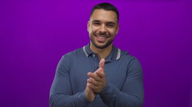 Mavi polo tişört giyen İspanyol genç adam, mor stüdyo ortamında geniş bir gülümsemeyle el çırpıyor;.