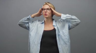 Beyaz tenli sarışın kadın ellerini şakaklara bastırır, gözlerini kapatır ve gri stüdyo arkaplanına yaslanırken yüzünü avuç içleriyle kapatır; stres baş ağrısı.