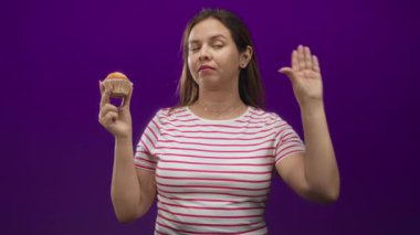 Mor bir stüdyoda bir eliyle kek tutarken diğer eliyle kulağını okşayan bir kadın..