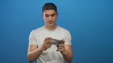 Latin asıllı genç adam elinde akıllı telefonla ekranı tıklatıyor, beyaz tişört giyiyor, mavi bir stüdyoda duruyor..