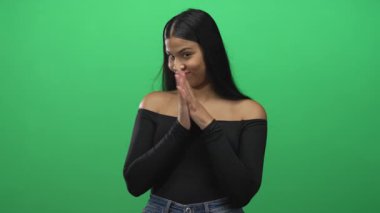 Genç Latin kadın, parlak yeşil bir stüdyo ortamında ellerini birbirine sürterek gülümser ve öne doğru eğilir..