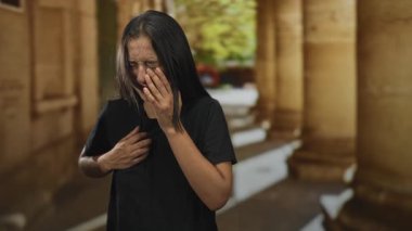 Yaşlı kasaba koridorunda kalp krizi geçirmek için göğsünü tutan genç İspanyol kadın elleri göğüs kafesine bastırıyor ve gözleri morarmış; panik içinde..