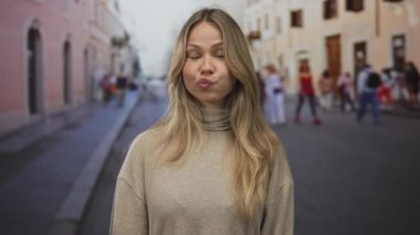 Sarışın kadın kaldırım taşı bir sokakta çenesinin altında kapalı el ele tutuşurken göz kırpar; şakacı merak neşe büyüsü.