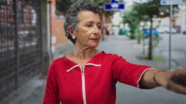 Son sınıf öğrencisi, gri saçlı, kırmızı elbiseli bir kadın gün boyunca şehir caddesini işaret ediyor..