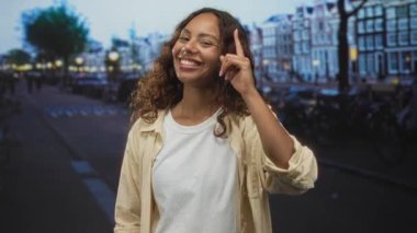 Kadın, beyaz tişörtlü ve açık gömlekli, görünür kolyeli, geniş bir gülümseme ile caddede yukarı işaret ediyor; mutluluk bağlantısı enerjisi.