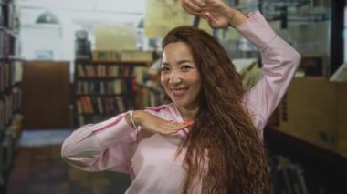 Elleri çerçeveli gülümseyen bir kadın kütüphane koridorunda kitapların arasında kolunu kaldırdı..