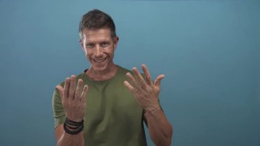 Avuç içlerini açan ve stüdyoda gülümseyen mavi arka plan ve günlük yeşil gömlekli bir adam. Dostça bir davet..
