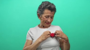 Gri saçlı yaşlı kadın yeşil arka planda kırmızı kalbi tutuyor, özen ve samimiyetini ifade ediyor..