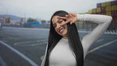 Genç İspanyol kadın gülümsüyor ve arka planda tekne ve deniz olan açık bir limanda gözleriyle barış jesti yapıyor..