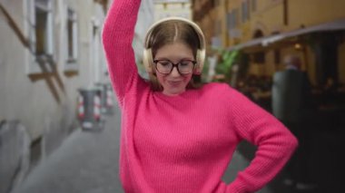 Young woman raises arm in pink sweater with white headphones on cobblestone street; joy freedom rhythm energy.