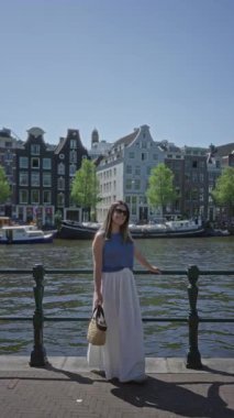 Geleneksel mimarisi ve arka planında tekneleri olan güneşli bir günde mavi bluz ve beyaz etek giyen, elinde saman torbasıyla gezen kadın..