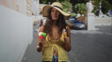 İki dondurma külahı tutan genç İspanyol kadın, gözleri kapalı ve gülümsüyor, hasır şapka ve örgü sarısı bluz giyiyor kaldırımlı bir sokakta dururken; yaz neşesi.