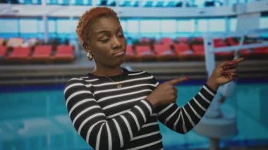 Kırmızı tırnaklı, çizgili siyah kazaklı kadın bina içindeki nesneyi işaret ediyor; şüphecilik.