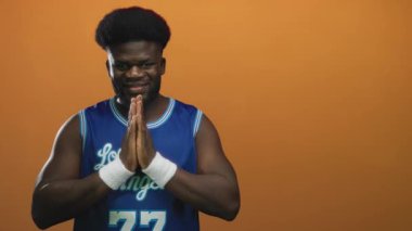 Elleri dua eden bir adam stüdyoda turuncu duvara karşı mavi Los Angeles basketbol forması giyiyor..