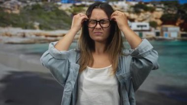 Gözlüklü kadın kumsalda kot gömlekli, beyaz kolsuz tişörtlü, görünür omuzlu ve yüzüklü ellerini kaldırıyor..