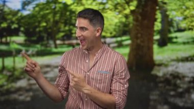 Yeşil ağaçların arasında parktaki iki parmağı gösteren adam, gülümseyen ve öne doğru eğilen; oyuncu özgüven.