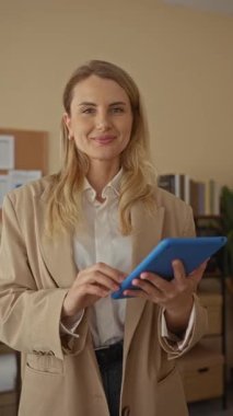 Ofiste elinde tablet olan genç bir kadın, kitaplık ve dosyalarla modern işyerinde güvenle gülümsüyor..