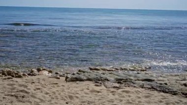 Akdeniz sahnesi berrak su, güneşli kum ve açık havada hafif dalgalar parlak mavi gökyüzünün altında sakin bir deniz atmosferini gösteriyor..