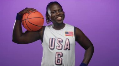 ABD forması ve bilekliğiyle mor stüdyoda basketbol tutan adam gülümsüyor ve poz veriyor; neşe atletizmi.