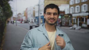 Kot ceket giyen bir adam, kalabalık bir şehir caddesinde saçlarına çıplak elle dokunurken gülümsüyor; kendine güven, soğukkanlılığını koruyor..